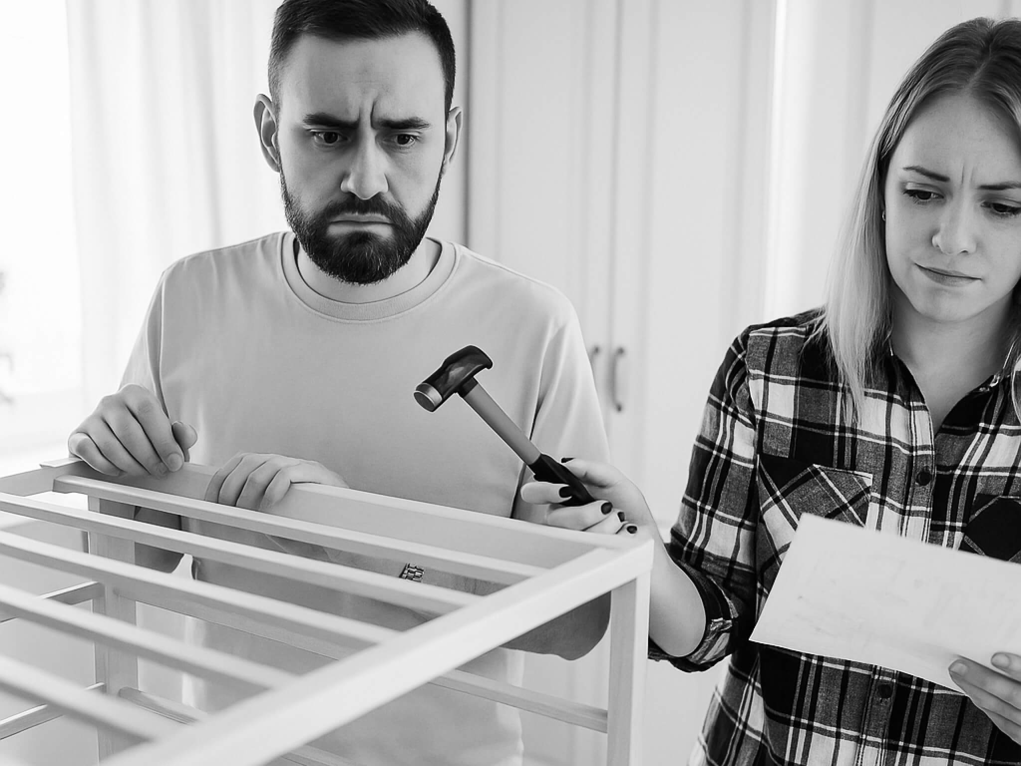 Una pareja con cara de frustración mientras monta una cuna, el hombre con un martillo y la mujer con unas instrucciones en papel poco claras.