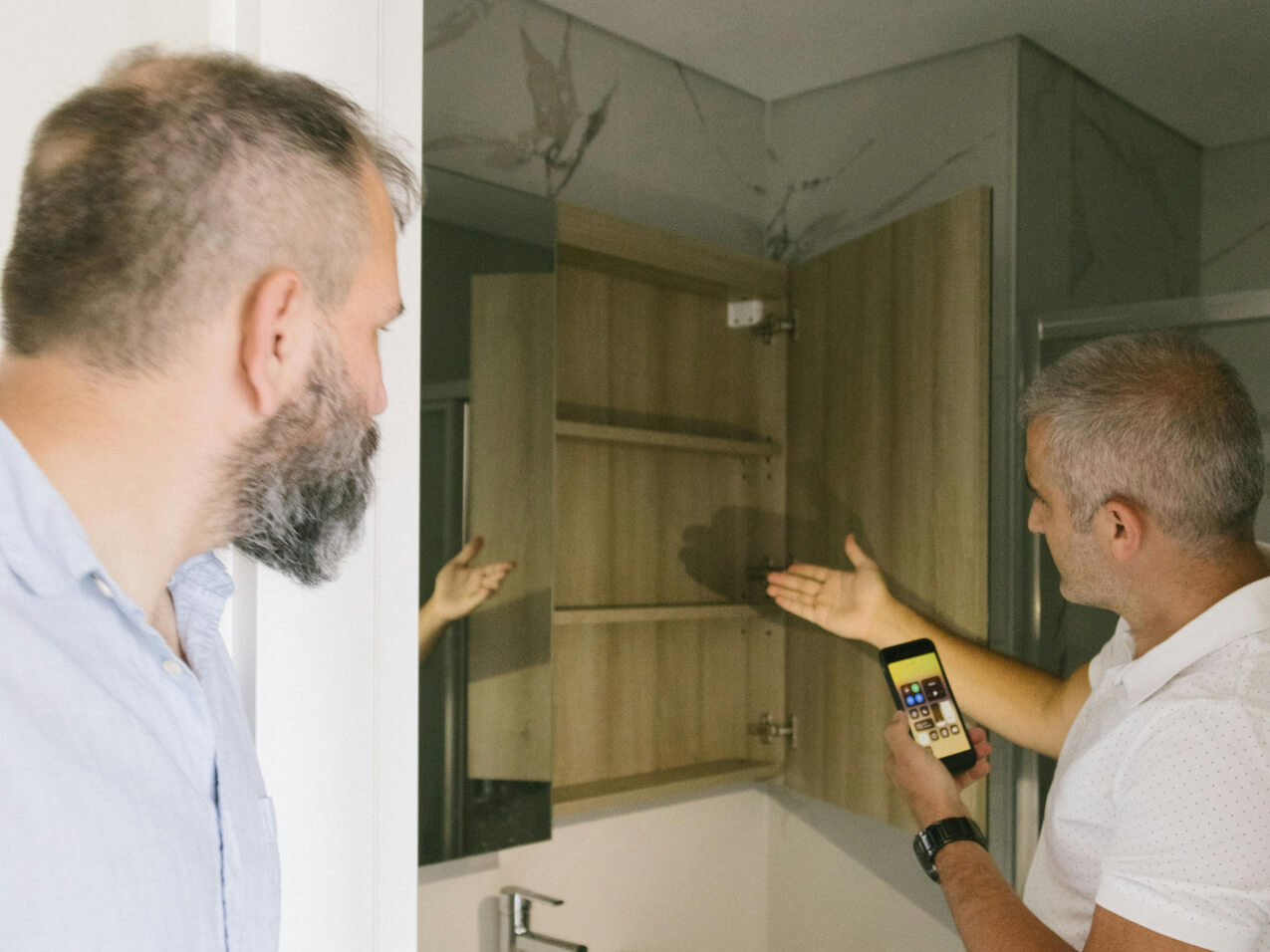 Dos hombres inspeccionan un mueble de baño mientras uno muestra comentarios en su teléfono.